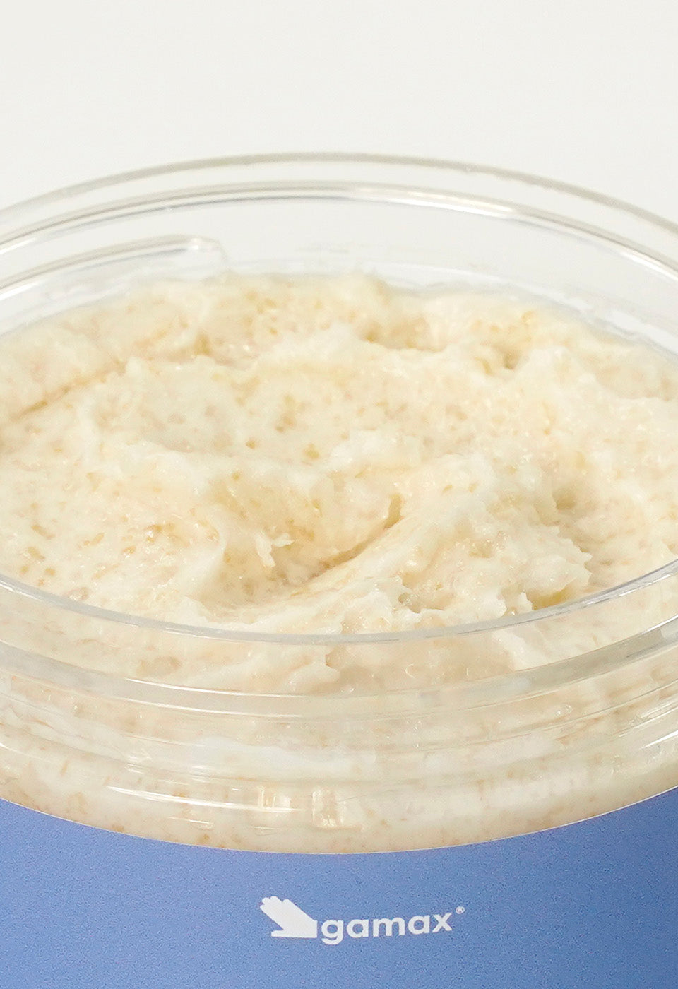 Dettaglio ravvicinato della texture dello scrub Gamax all'interno del barattolo. La consistenza appare come un'emulsione densa e ricca di granuli esfolianti color crema, ideale per levigare la pelle.