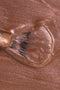 Dettaglio ravvicinato della texture di uno smalto color bronzo rosato metallizzato. Un pennellino con setole nere è immortalato mentre stende il prodotto, creando venature lucide e micro-glitterate sulla superficie