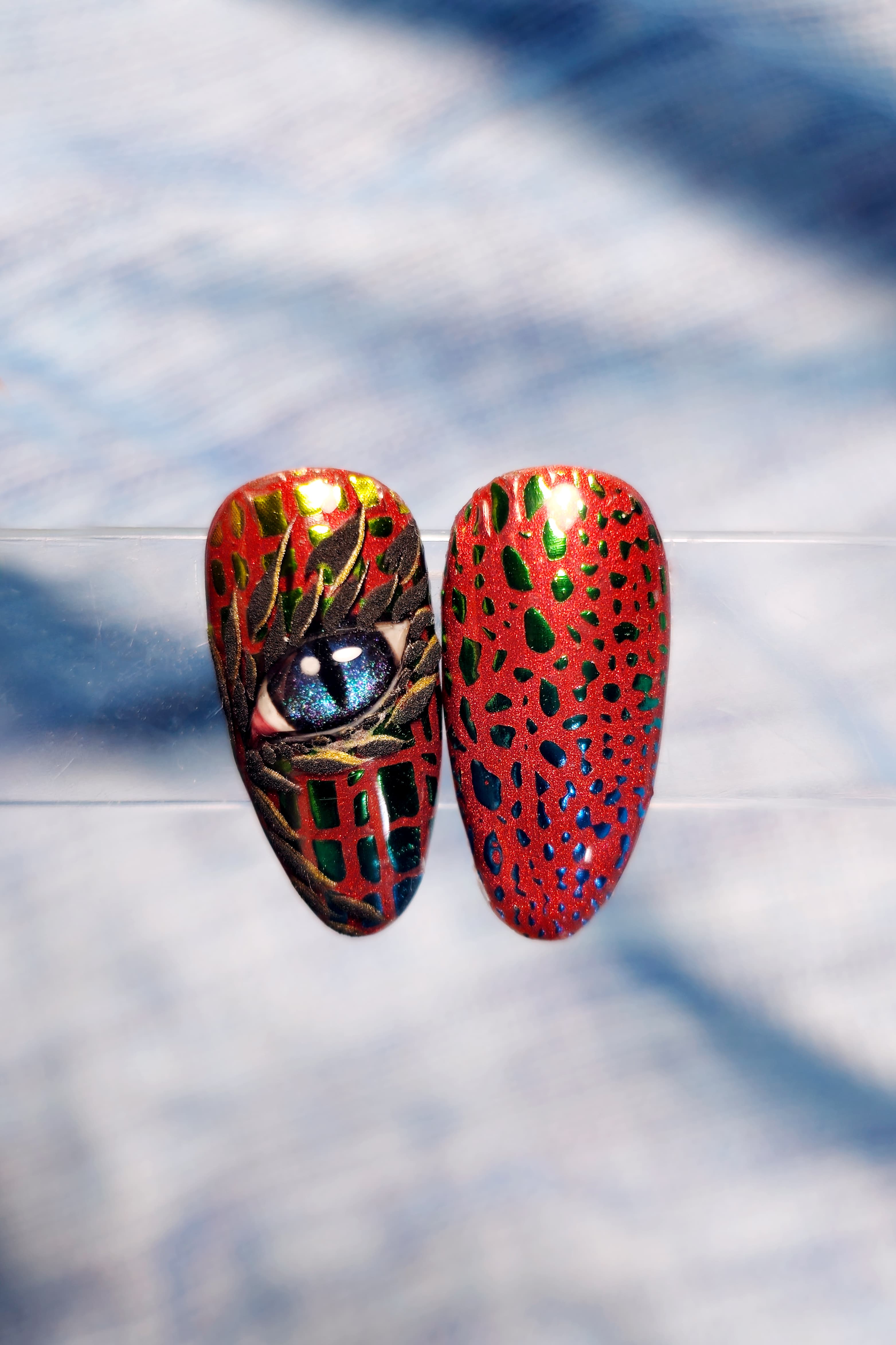 Nail Art su tip realizzata con adesivi fantasia squame di drago arcobaleno. Base rossa, adesivi squama di drago arcobaleno iridescenti. La tip a sinistra ha una nail art realizzata a mano raffigurante un occhio di drago azzurro.