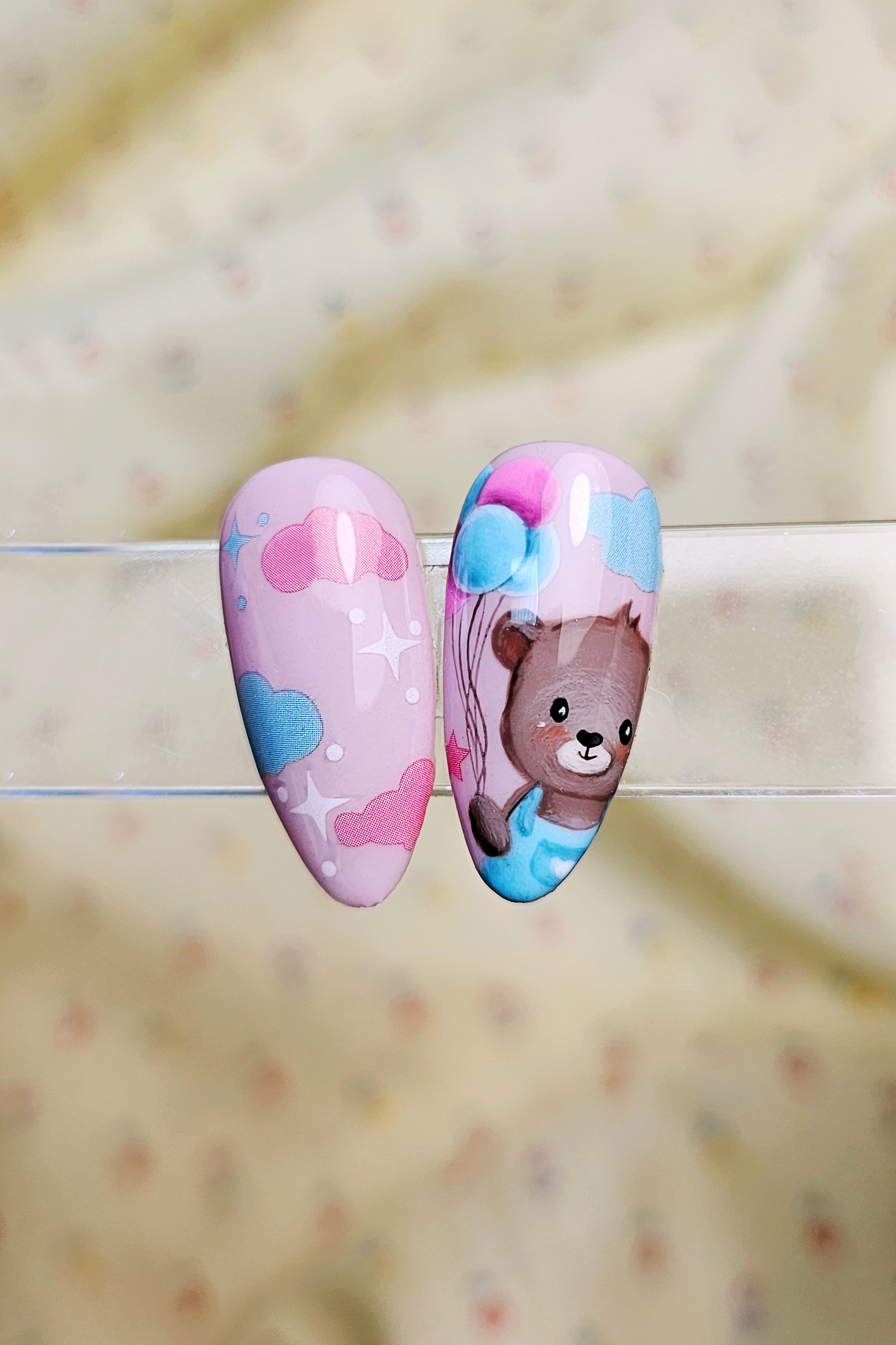 Nail Art su due tip. Base rosa pastello. Realizzato con sticker unghie di nuvole colorate e stelline bianche colorata. Il soggetto principale è un orsacchiotto marrone con maglia azzurra che tiene dei palloncini.