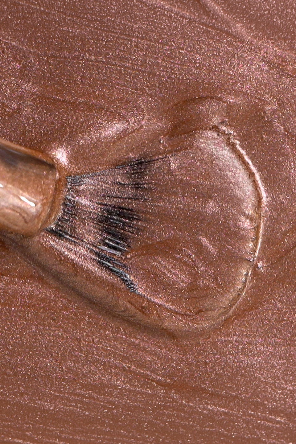 Dettaglio ravvicinato della texture di uno smalto color bronzo rosato metallizzato. Un pennellino con setole nere è immortalato mentre stende il prodotto, creando venature lucide e micro-glitterate sulla superficie