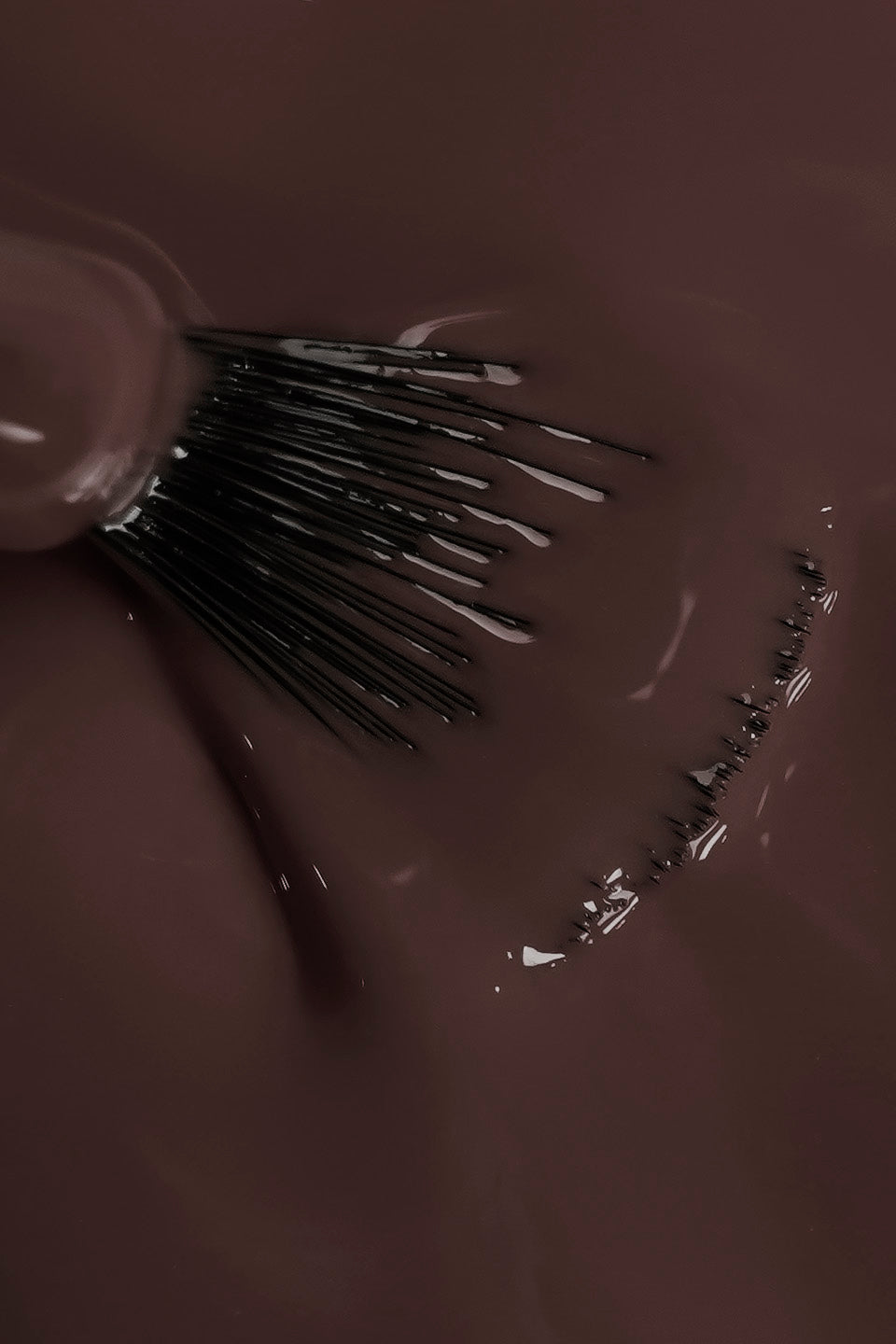 Dettaglio ravvicinato della texture fluida dello smalto semipermanente marrone scuro ambra. Un pennello a ventaglio è immerso nel colore, mostrando la coprenza omogenea e la finitura lucida del prodotto durante la stesura.