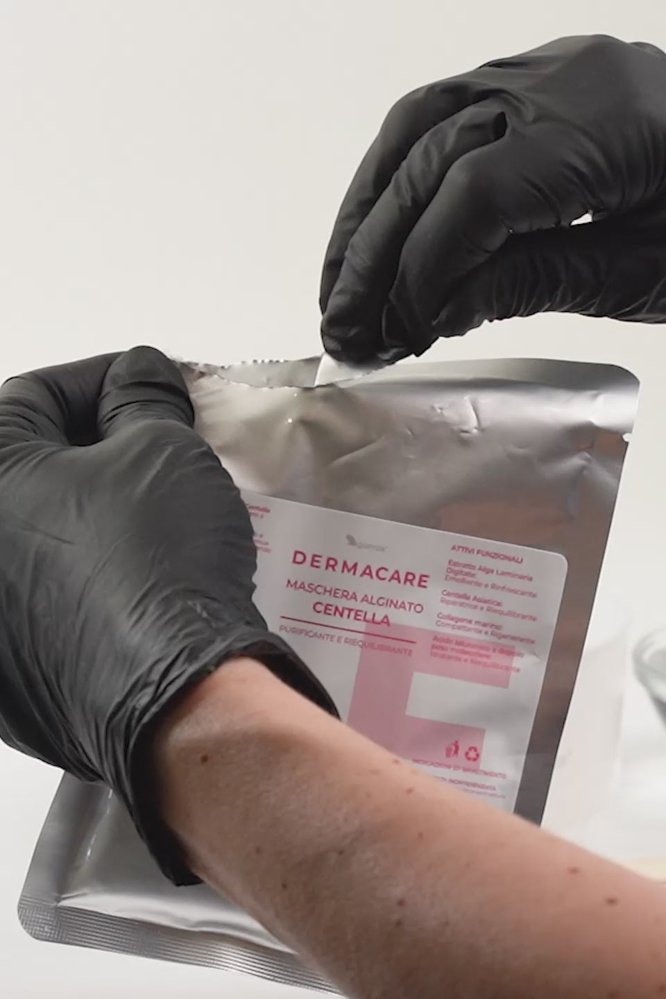 Video che mostra la preparazione e l'applicazione di una maschera viso professionale. Una persona con guanti neri apre una busta metallica con l'etichetta "DERMACARE MASCHERA ALGINATO CENTELLA" e versa una polvere verde chiaro con frammenti botanici in una ciotola bianca. Dopo aver aggiunto dell'acqua, mescola il composto con una spatola di legno fino a ottenere una consistenza gelatinosa e uniforme. La maschera viene poi stesa delicatamente sul viso  e, una volta solidificata, viene rimossa.