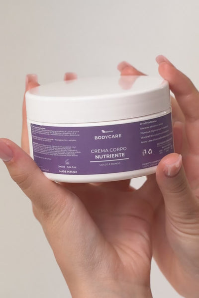 Primo piano ravvicinato dell'applicazione di una crema corpo. All'inizio del video, una persona mostra un barattolo bianco con l'etichetta viola che recita "BODYCARE - CREMA CORPO NUTRIENTE - COCCO E MANGO". Subito dopo, la scena passa al dorso di una mano su cui è stata versata una generosa quantità di crema bianca; delle dita con un'elegante manicure rosa chiaro sfumano e massaggiano delicatamente il prodotto sulla pelle, evidenziandone la consistenza ricca e idratante.
