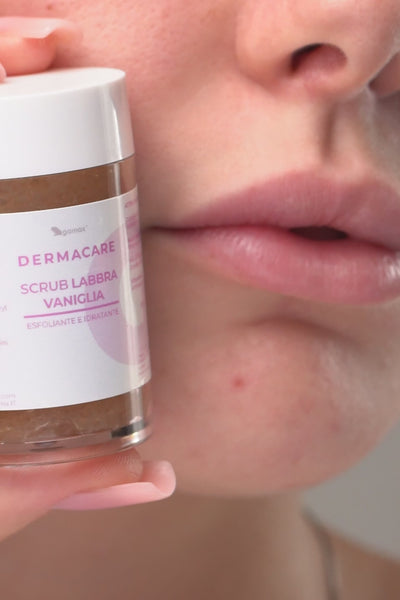 Primo piano ravvicinato dell'applicazione di uno scrub labbra. Il video mostra una persona che tiene in mano un barattolo di prodotto con l'etichetta "DERMACARE SCRUB LABBRA VANIGLIA". Successivamente, una spatola di legno applica il composto esfoliante, ricco di granuli di zucchero dorati, direttamente sulle labbra. Infine, un dito con manicure rosa massaggia delicatamente il prodotto sulle labbra per completare l'esfoliazione.