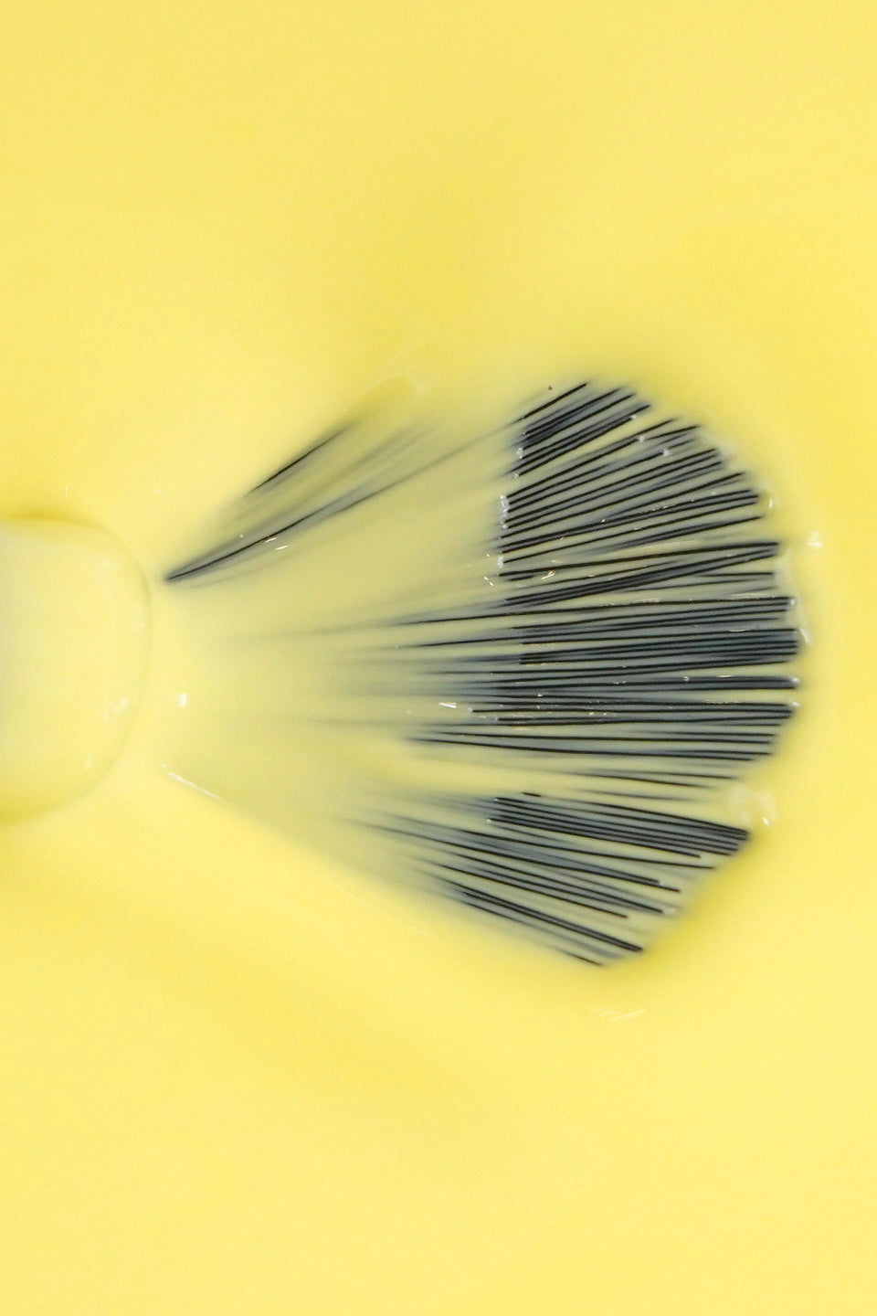 Inquadratura ravvicinata dall'alto che mostra la texture di uno smalto liquido color giallo chiaro. Al centro, le setole nere di un pennellino da stesura sono aperte a ventaglio e parzialmente immerse nel prodotto, evidenziando la consistenza fluida, cremosa e coprente dello smalto.