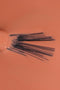 Inquadratura ravvicinata dall'alto che mostra la texture di uno smalto liquido color arancione chiaro pesca. Al centro, le setole nere di un pennellino da stesura sono aperte a ventaglio e parzialmente immerse nel prodotto, evidenziando la consistenza fluida, cremosa e coprente dello smalto.
