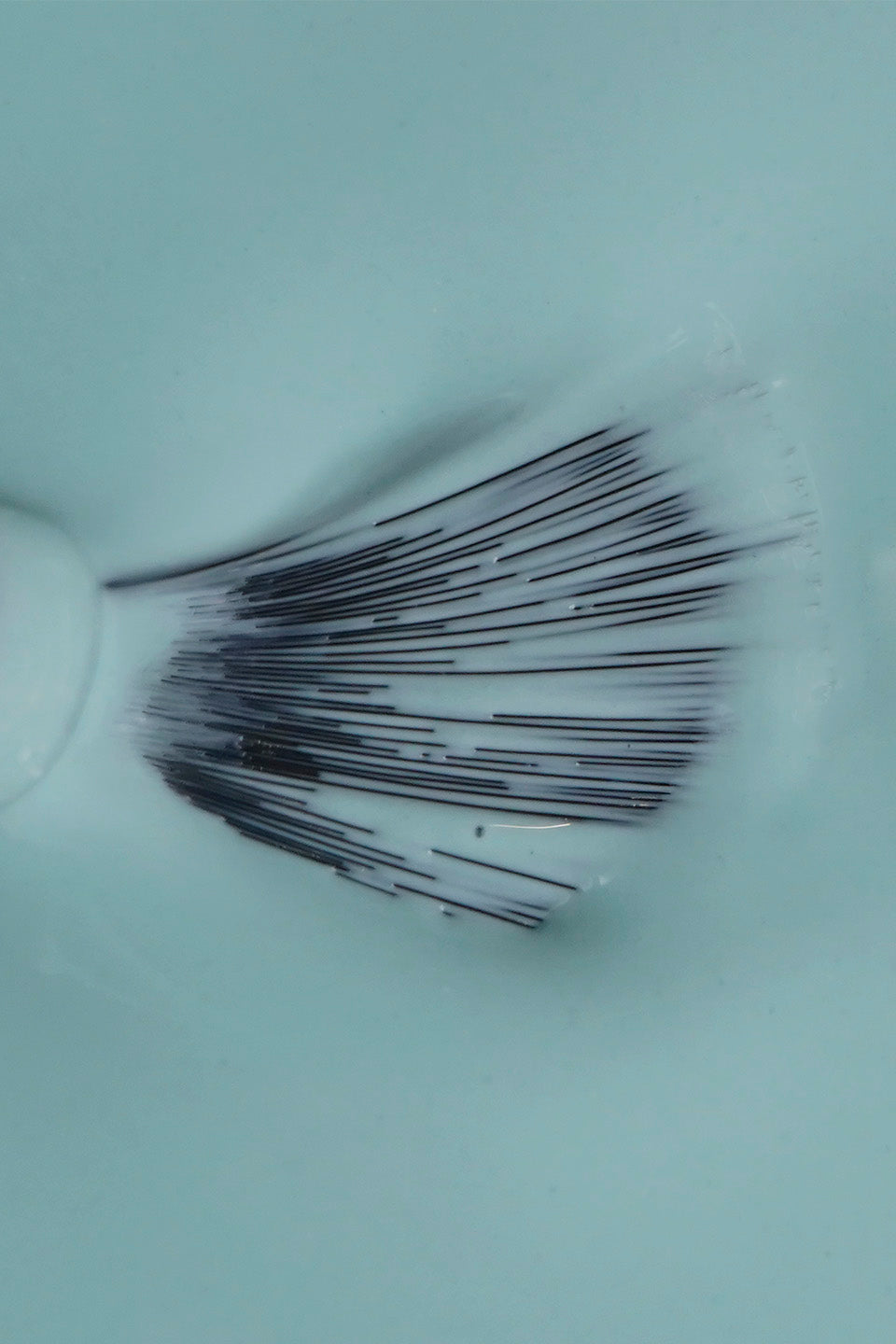 Inquadratura ravvicinata dall'alto che mostra la texture di uno smalto liquido color turchese chiaro. Al centro, le setole nere di un pennellino da stesura sono aperte a ventaglio e parzialmente immerse nel prodotto, evidenziando la consistenza fluida, cremosa e coprente dello smalto.